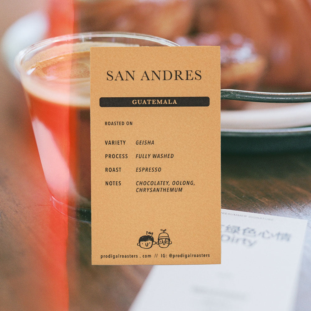 A vertical kraft paper card with black text reading 'SAN ANDRES' and 'GUATEMALA' at the top, listing roast and flavor notes below, photographed on a wooden table with a blurred coffee cup in the background.